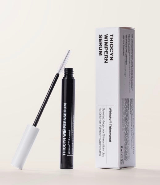 In der Mitte steht das geöffnete Thiocyn Wimpernserum, links daneben lehnt die Wimpernbürste an die Flasche des Wimpernserums und rechts daneben steht die Umverpackung des Wimpernserums. In der Mitte steht das geöffnete Thiocyn Wimpernserum, links daneben lehnt die Wimpernbürste an die Flasche des Wimpernserums und rechts daneben steht die Umverpackung des Wimpernserums.