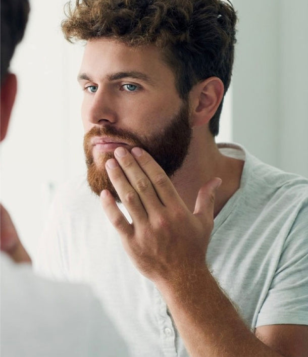 Braunhaariger Mann streicht sich durch seinen bart und betrachtet sich dabei im Spiegel.